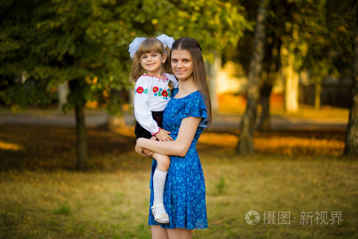 秋天公园背景上美丽的年轻母亲抱着一年级的女儿穿着节日校服的肖像