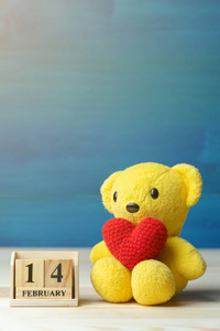 手工制作纱线红心放在黄色泰迪熊旁边的木块日历上，日期为2月14日，木制桌子和蓝色背景。 情人节的概念。