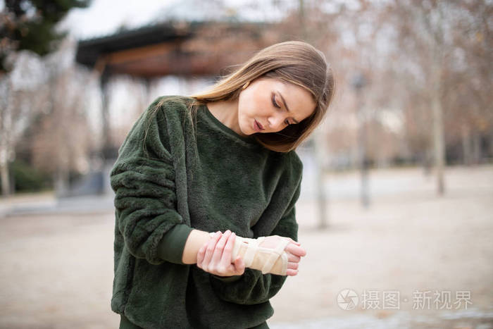 美丽的年轻女士穿着时尚的服装皱眉抚摸受伤的手臂上的绷带站在秋天的