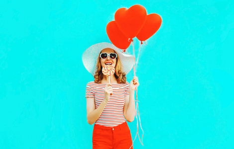 快乐微笑的年轻女子，带着棒棒糖，红色心形气球，戴着夏草帽，穿着五颜六色的蓝色背景短裤