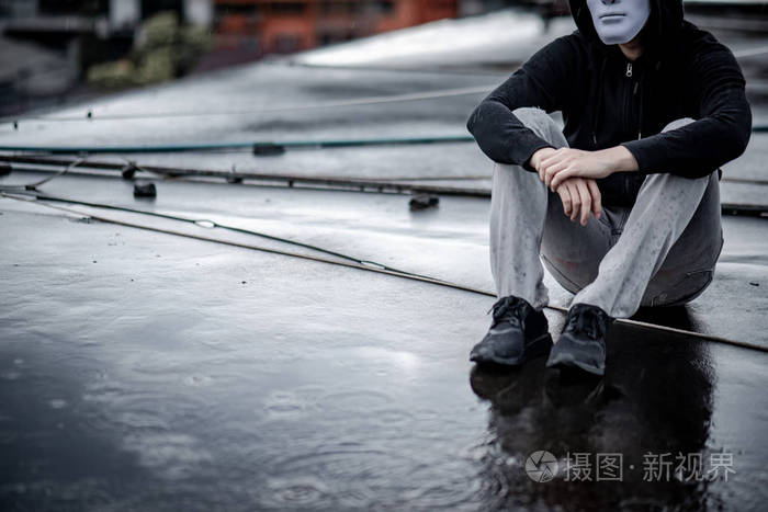 神秘的帽衫男人在白色面具下拥抱着他的膝盖,坐在废弃建筑的屋顶上的