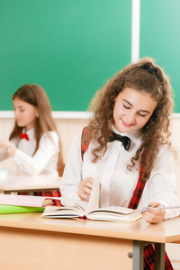 两个女学生坐在教室里的桌子上上课。 一个女孩坐在一张桌子旁，拿着书和学习。女学生在教室里坐在桌子旁思考