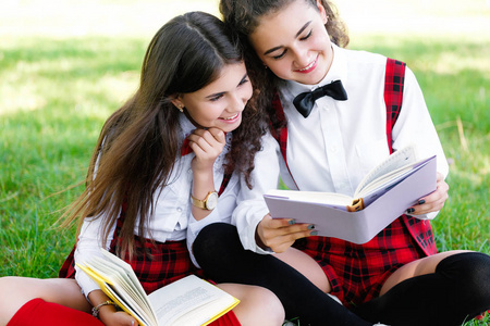 两个穿红色校服的女学生坐在绿色的草坪上，带着书。女朋友或姐妹教户外课程