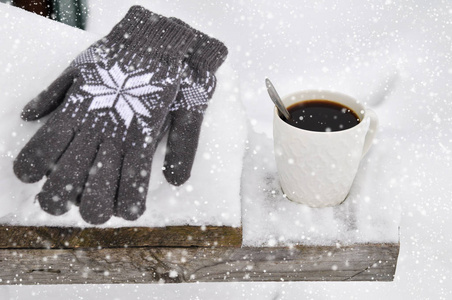 一杯白色的咖啡和针织的灰色手套，在降雪时雪地里的木凳上有图案。 舒适的冬卡。