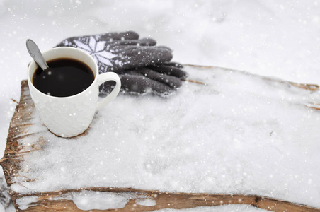 一杯白色的咖啡和针织的灰色手套，在降雪时雪地里的木凳上有图案。 舒适的冬卡。
