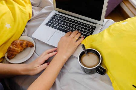女人的手和笔记本，牛角面包和咖啡在床上用黄色的粉彩。 顶部视图