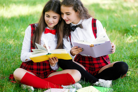两个穿红色校服的女学生坐在绿色的草坪上，带着书。女朋友或姐妹教户外课程