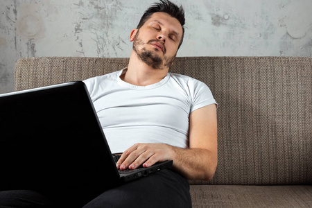 坐在沙发上的那个穿白色衬衫的家伙在笔记本电脑上工作时睡着了。 懒惰冷漠挫折的概念拖延了家里的人。 复制空间。