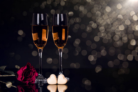两杯香槟玫瑰和两颗心形巧克力在黑色背景与灯光。 爱情人节的概念。 复制空间