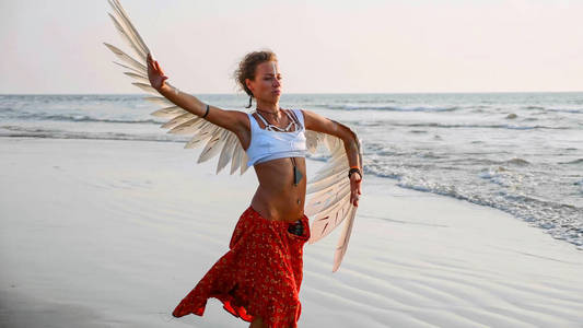在日落时在沙滩上跳舞的年轻女孩与天使的翅膀
