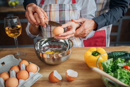 男子双手帮助妇女用刀打破鸡蛋。他站在她身后的厨房里。桌子上有沙拉配料和玻璃