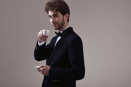 时尚肖像，英俊的优雅男人，卷发，穿着燕尾服，在工作室的灰色背景上拿着一杯浓缩咖啡