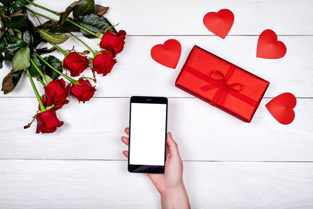 手机空白屏幕在女人手中，玫瑰花束，心和礼品盒在白色背景复制空间。 平躺顶部视图。 情人节女人日母亲日