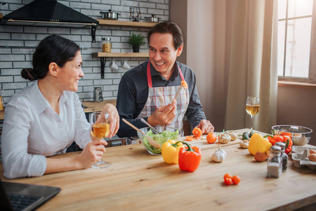快乐的男人和妻子坐在厨房里的桌旁。他把一块西红柿放在叉子上给女人看。她看着它, 微笑着。peoepl 准备晚餐和喝葡萄酒
