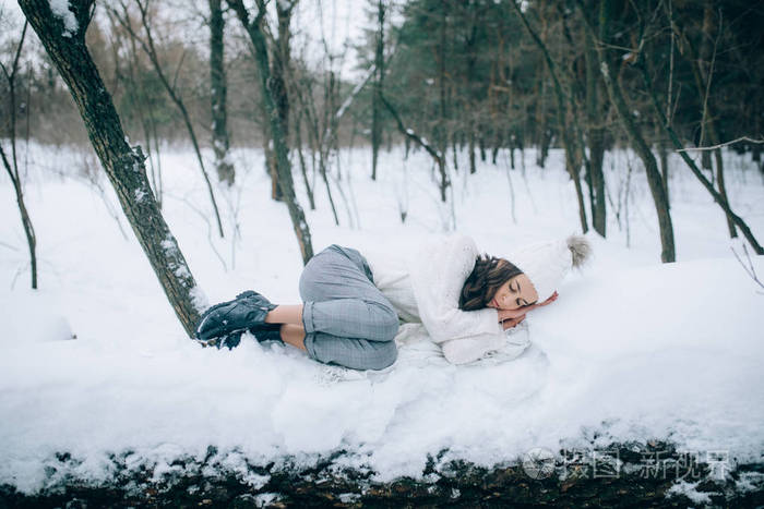 在冬天的森林里散步时年轻悲伤的女人躺在白雪覆盖的树干上休息