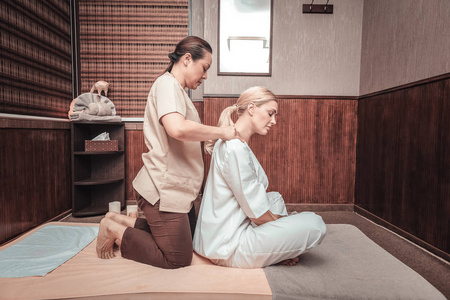 美丽的金发女人坐在按摩师前面,一边享受肩部按摩照片