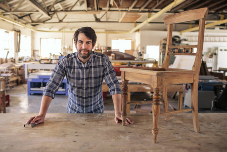 一个微笑的家具制造商的肖像靠在他的木工工作室的长凳上，同时打磨一把椅子