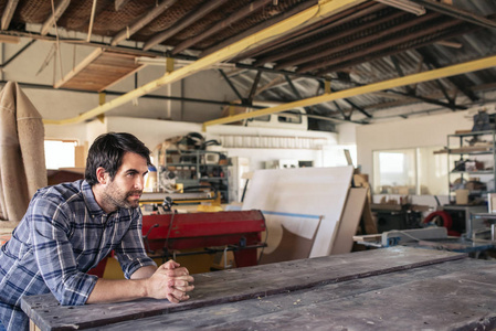年轻的木工在他的大型家具制作工作室里靠在一个工作台上，一边深入思考