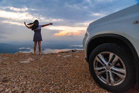 女人站在汽车附近的悬崖上，日出时看到美丽的湖景。公路旅行