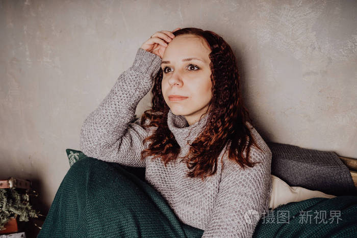 一个穿毛衣的年轻女孩坐在床上,蜷缩在毯子里.