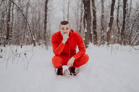 在冬天的森林里，在雪和树的背景下，疯狂的家伙在摆姿势。 一个穿着红色运动服在新鲜空气中行走的人。 概念在寒冷的户外散步和训练