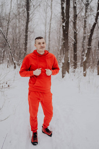 在冬天的森林里，在雪和树的背景下，疯狂的家伙在摆姿势。 一个穿着红色运动服在新鲜空气中行走的人。 概念在寒冷的户外散步和训练