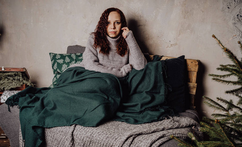 一个穿着毛衣坐在床上的年轻女孩蜷缩在毯子里.