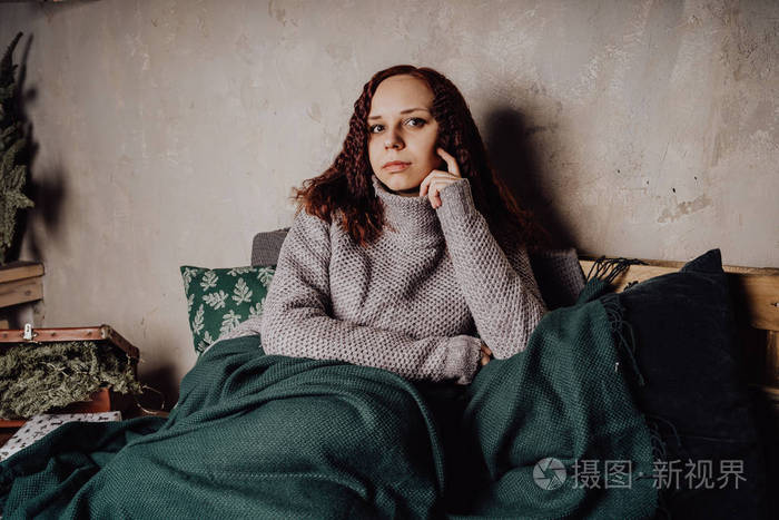 一个穿着毛衣坐在床上的年轻女孩蜷缩在毯子里.