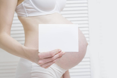 孕妇拿着一张白色的白纸。 肚子在背景上。 怀孕概念