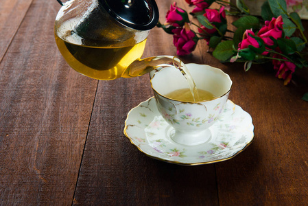 用粉红色的花从木制桌面上的玻璃壶中倒入一杯茶