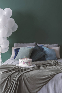 生日蛋糕上有蜡烛，舒适的双人床上有枕头，空的绿色墙上有毯子，角落里有一堆白色气球