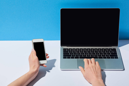 使用带有空白屏幕的笔记本电脑和白色桌子和蓝色背景上的智能手机的女性裁剪视图