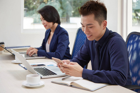 微笑的年轻商人坐在桌子旁，在工作日用手机发短信，同事在后台用笔记本电脑打字