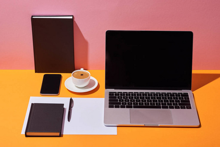 笔记本电脑，带有空白屏幕，靠近咖啡杯茶托笔纸和黄色书桌和粉红色背景的笔记本电脑