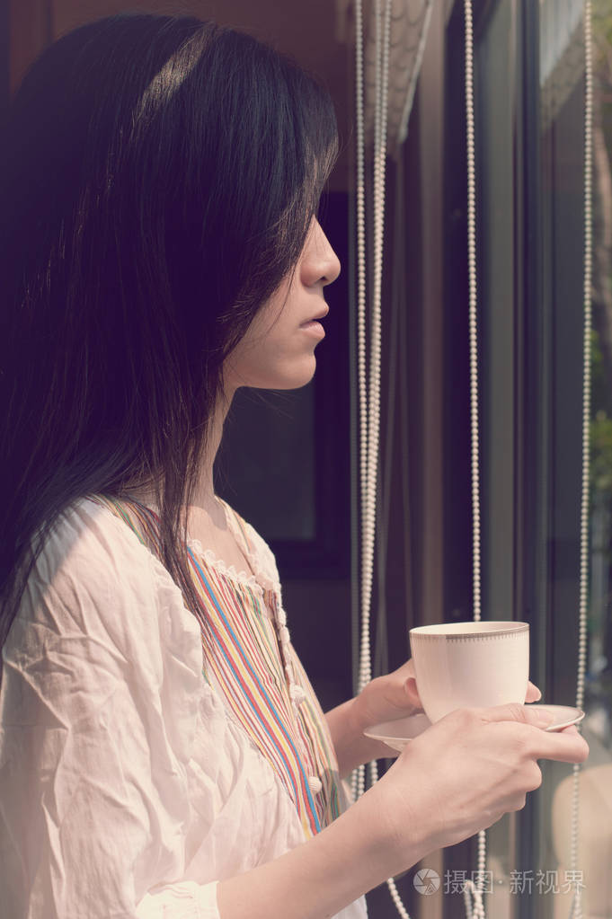 亚洲女人看起来忧郁和孤独,透过窗户,一杯咖啡或茶的复古风格.