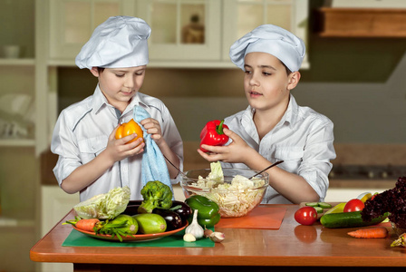 穿着厨师服装的男孩在厨房切沙拉。 孩子们准备健康的食物。 厨师的友好合作。 那个男孩用毛巾擦蔬菜。