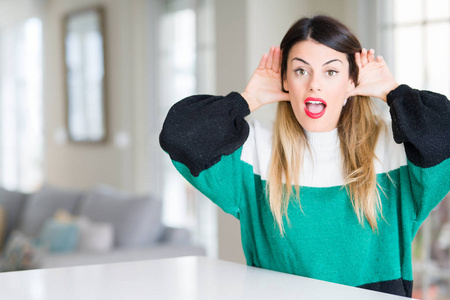 年轻漂亮的女人穿着冬天的毛衣在家里，试图听到双手在耳朵上的手势好奇八卦。 听力问题