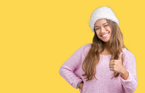 年轻漂亮的黑发女人，穿着毛衣和冬帽，在孤立的背景上，用手做快乐的竖起大拇指的手势。 赞许的表情看着相机显示成功。