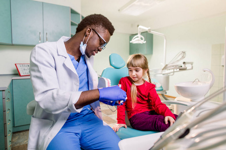 小女孩坐在牙科椅上，手里拿着人造下巴对着非裔美国人牙医微笑
