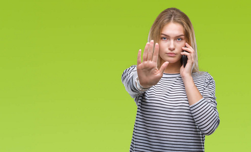 年轻的白种人妇女在孤立的背景上显示智能手机屏幕，张开手做停止标志，严肃和自信的表情防御手势。