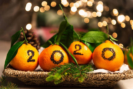 新年2020即将到来的概念。 用黑色墨水写在篮子里的橘子上的数字，背景上有松枝和圣诞灯