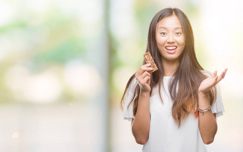 年轻的亚洲妇女吃巧克力，精力充沛的酒吧，在孤立的背景下，非常高兴和兴奋的胜利者的表情，庆祝胜利，微笑着尖叫，举起双手