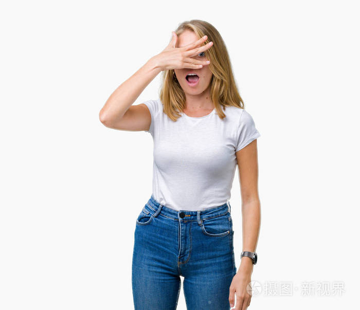 美丽的年轻女子穿着休闲的白色t恤在孤立的背景上偷看震惊地遮住脸和