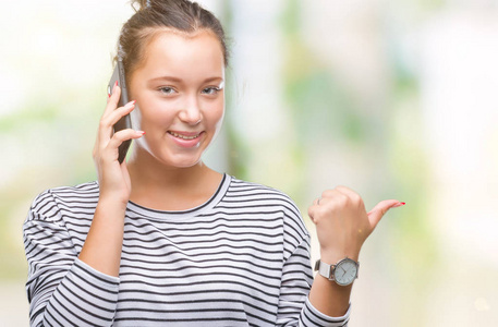 年轻美丽的白种人女人在智能手机上交谈，在孤立的背景下，指着并用拇指向侧面展示，脸上洋溢着幸福的微笑