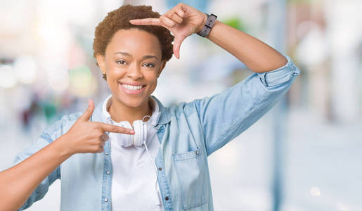 年轻的非裔美国妇女戴着耳机，在孤立的背景上微笑，用手和手指制作框架，用快乐的脸。 创造力和摄影理念。