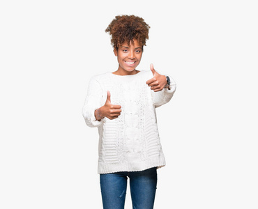美丽的年轻非洲裔美国妇女穿着冬季毛衣，在孤立的背景下，批准用手竖起大拇指做积极的手势，微笑，为成功而高兴。 看着相机赢家的手势。