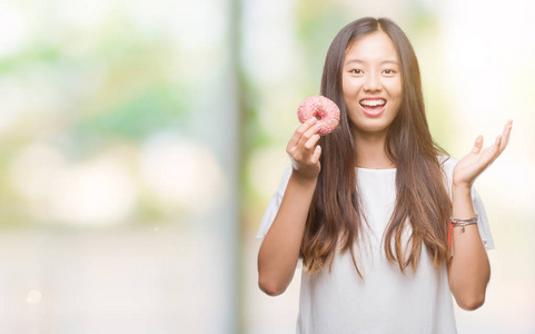 年轻的亚洲女人在孤立的背景下吃甜甜圈，非常高兴和兴奋，胜利者的表情庆祝胜利，大笑着尖叫，举手