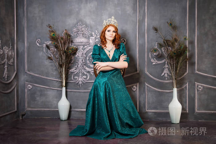 年轻优雅的女人穿着祖母绿的衣服戴着豪华的钻石皇冠坐在灰色的内部的