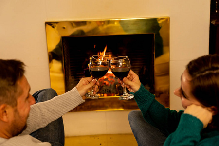 亲密的浪漫夫妇举着两个红酒杯在火中品尝，在寒假假期圣诞节或周年纪念的概念中，一起在小屋的壁炉旁庆祝快乐的时刻。