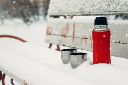 情侣的冬季约会想法。 寒假热饮概念。 冬天公园雪凳上的红色热水瓶和杯子
